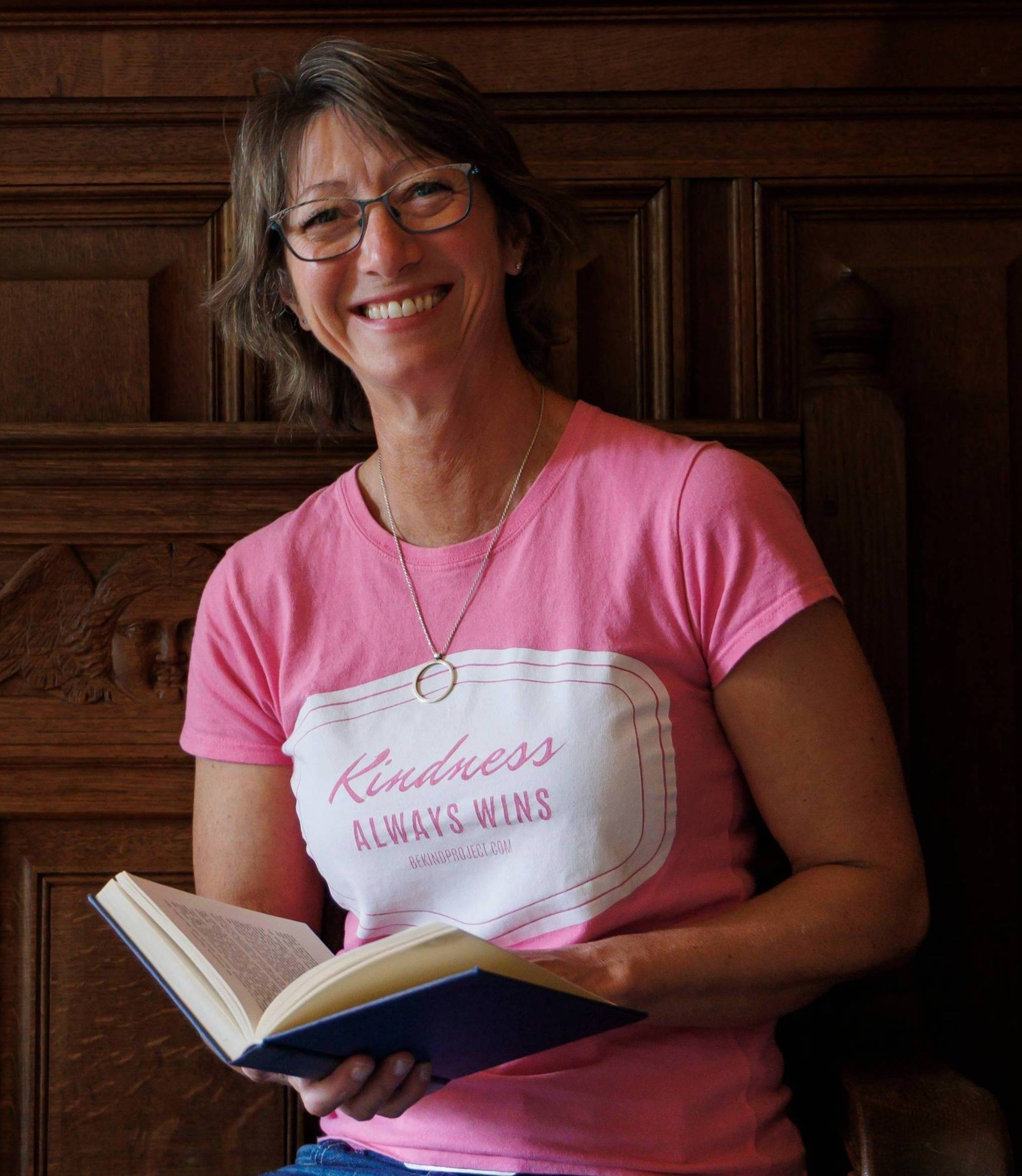 Woman in a pink t-shirt holds a book and smiles at the camera.