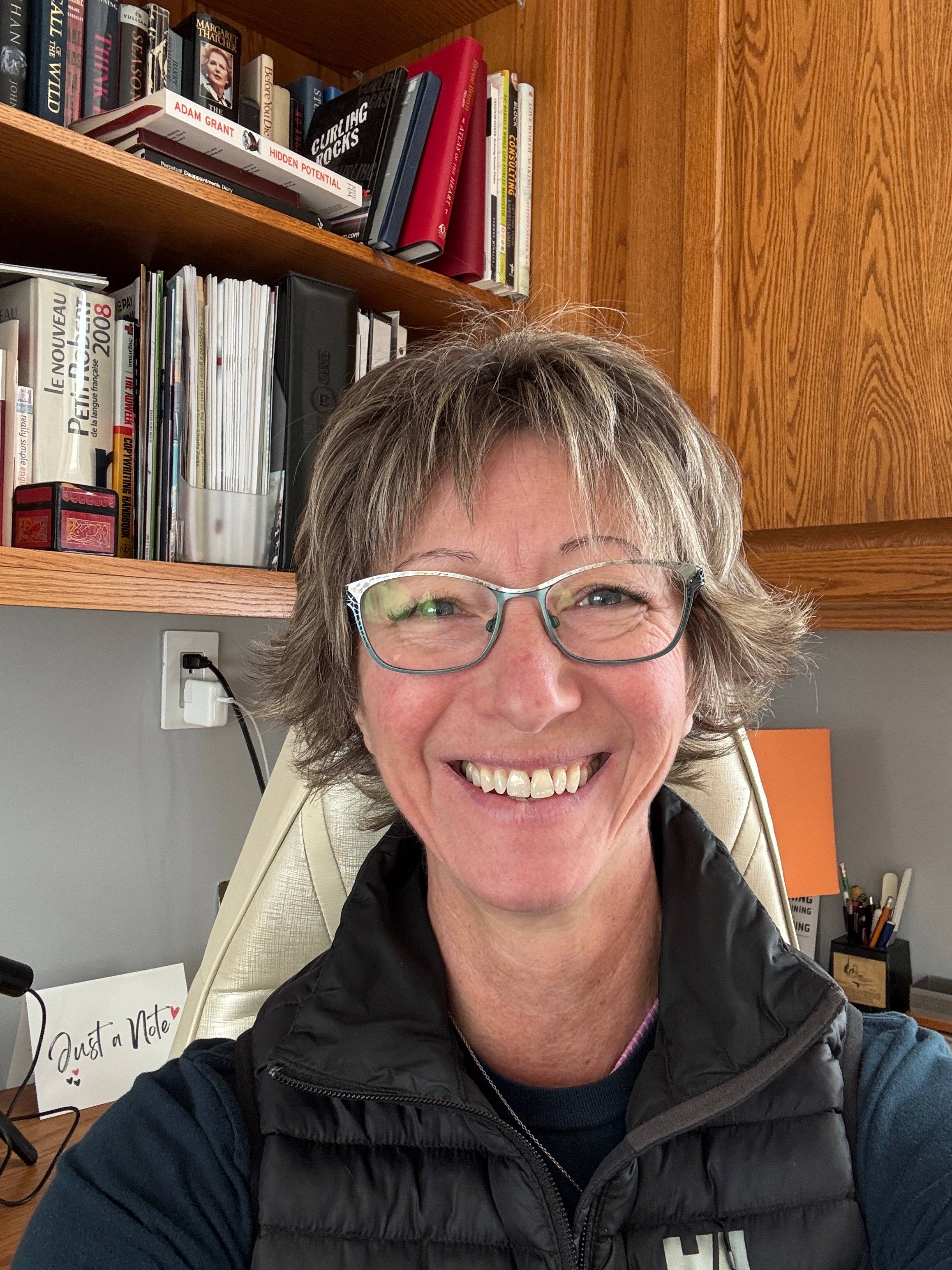 A woman with glasses and short blond hair smiles into the camera.