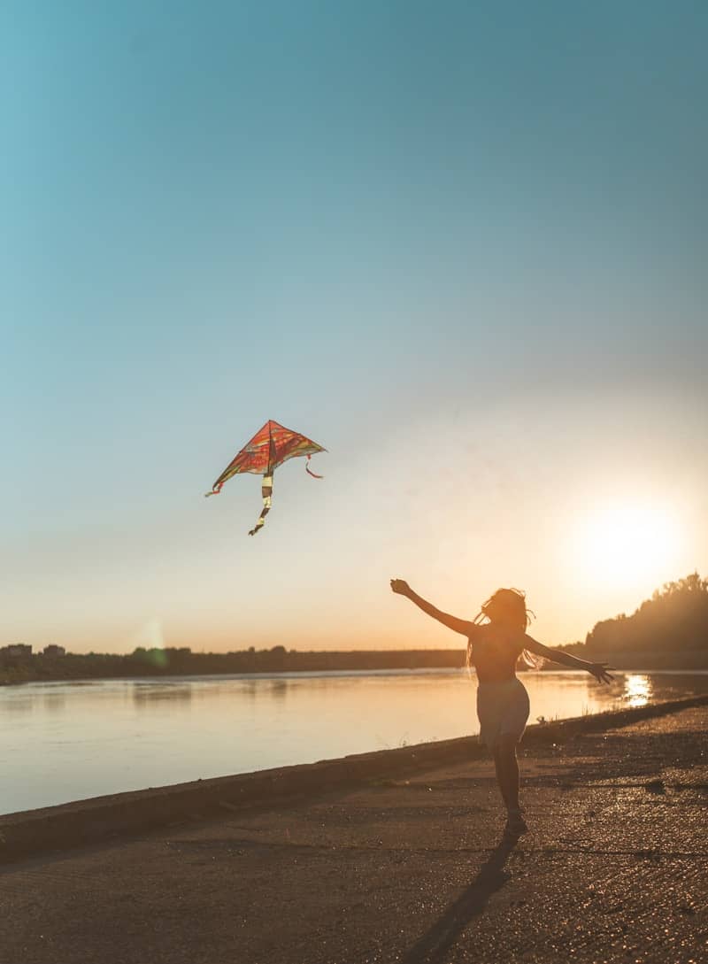 A person in silhouette running with a colorful kite by a river at low sun.