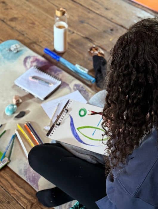 Women with curly brown hair seated on ground coloring on paper. 