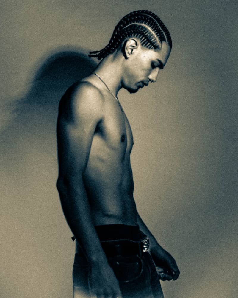 Man with cornrows looking down against a wall.