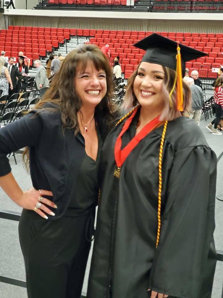 Two women stand smiling next to each other, one is wearing a graduation cap and robes. They are mother and daughter.