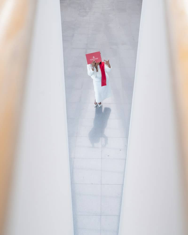 A person in a white suit holding a red folder over their face in a bright hallway