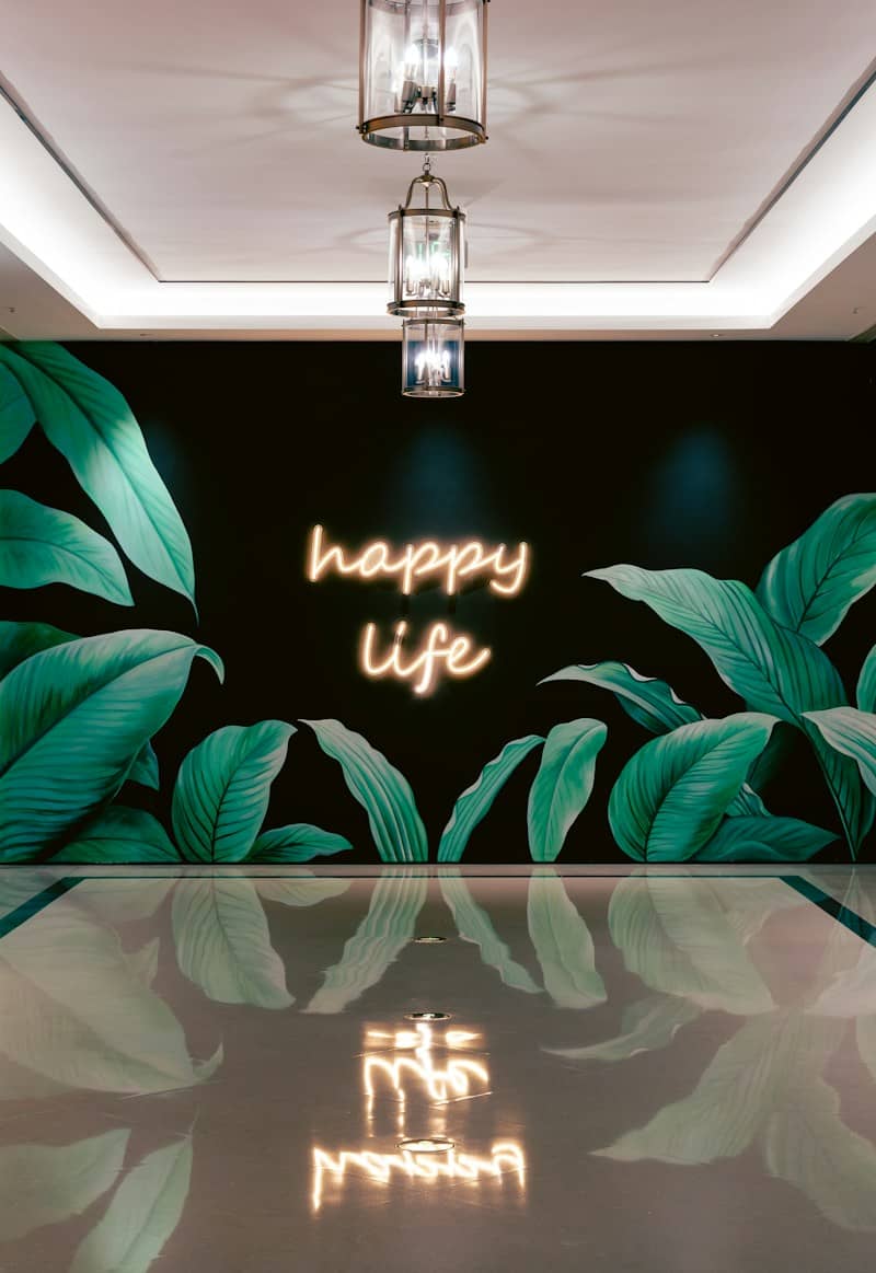 A neon sign reading "happy life" on a black wall with painted green leaves