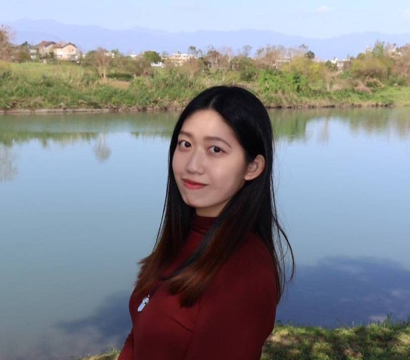 A woman with long black hair and a maroon top is standing near a calm body of water. The background features lush green vegetation and distant mountains under a clear blue sky.