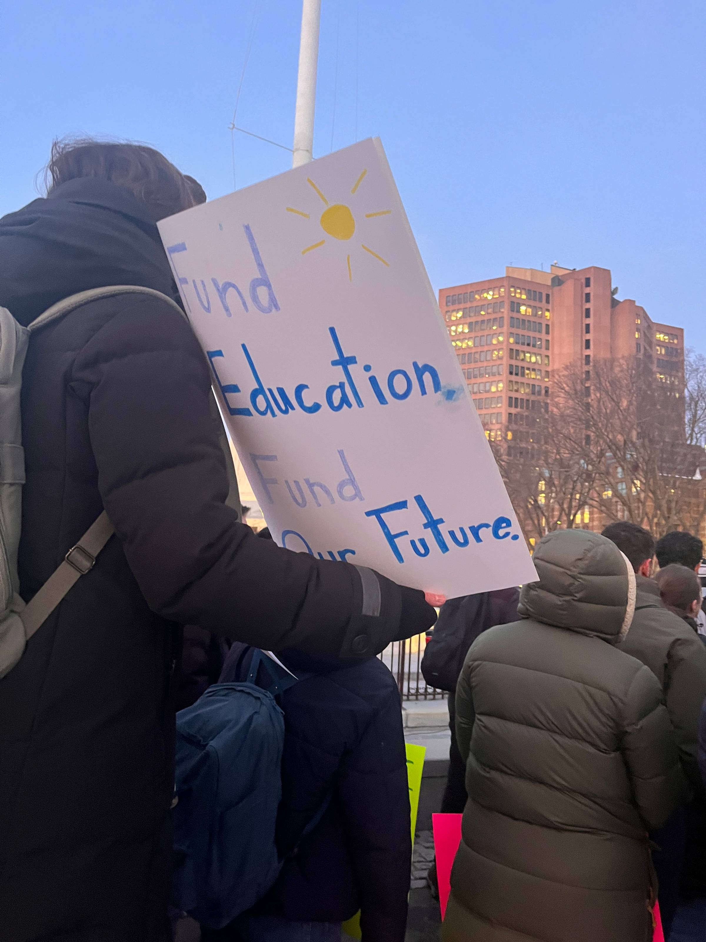 A person bundled up in a parka witha sign that reads "fund education, fund our futures" with city buildings in the distance