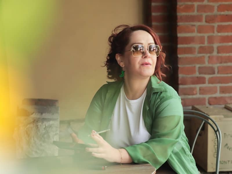 A woman with red hair wearing sunglasses and a green shirt holding a cigarette