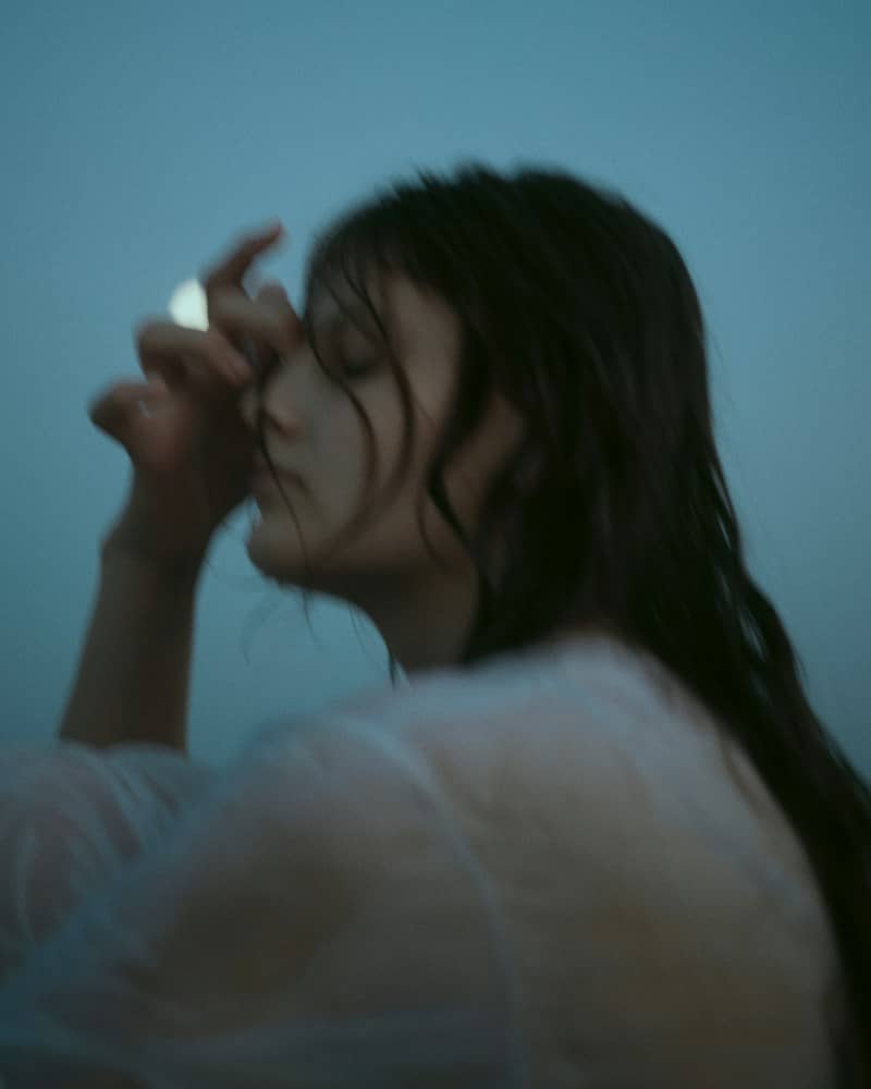 A person with long dark hair touching their forehead against a blue background