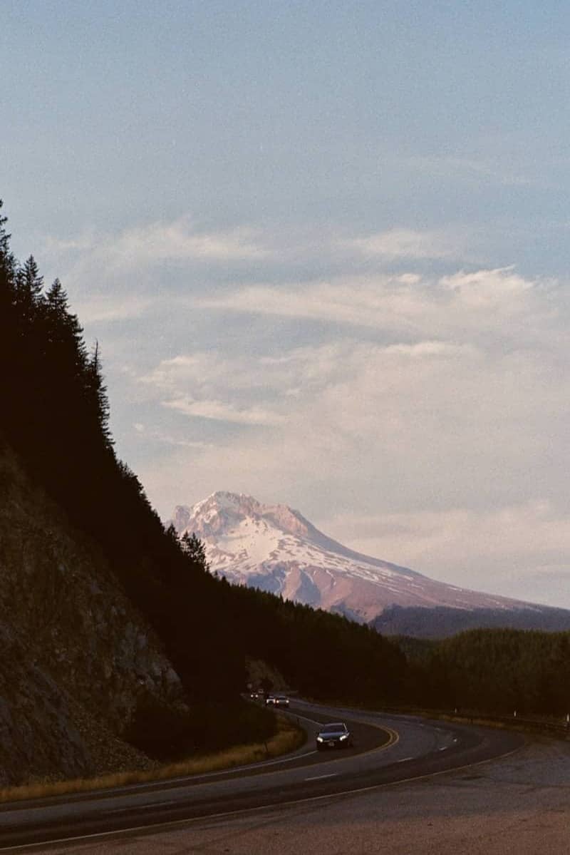 Cars drive on a winding road towards a snow-capped mountain.