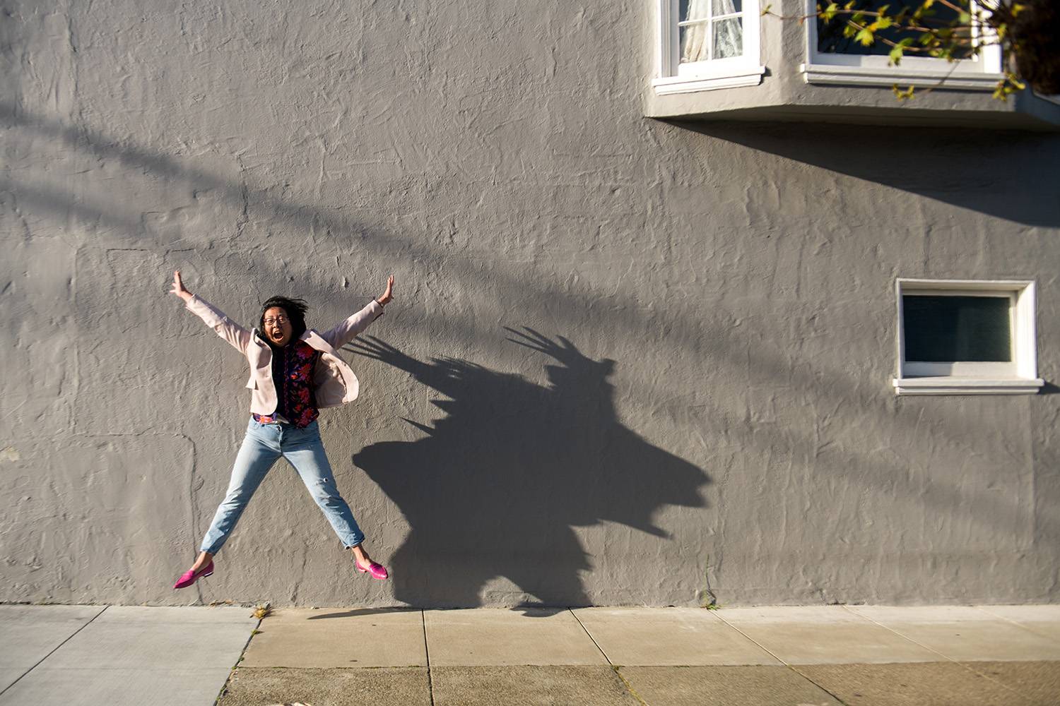 A woman leaping in front of a gray wall, her long shadow stretching beside her