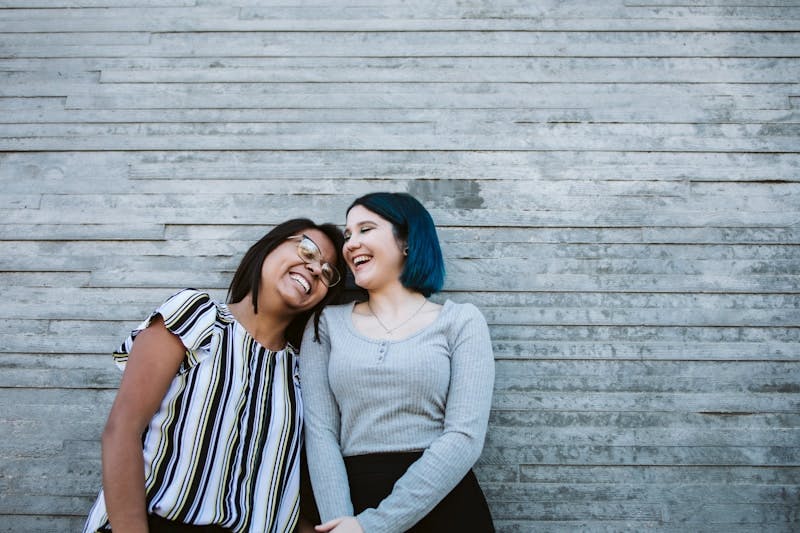 2 women smiling and standing beside gray wooden wall