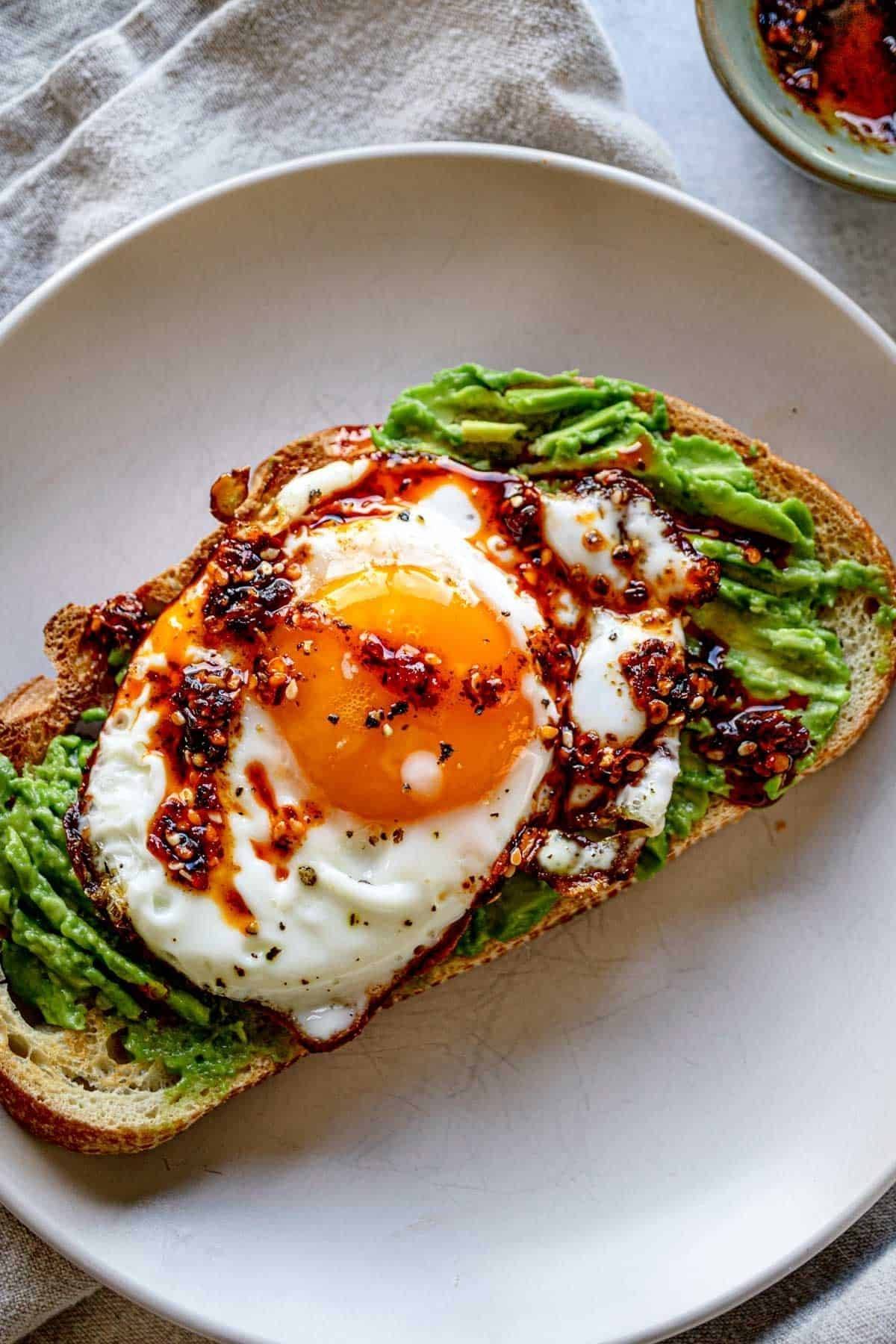 A plate with a slice of toast topped with mashed avocado, a fried egg, and a drizzle of chili oil. The yolk is bright and slightly runny, with a sprinkle of black pepper on top. A napkin and a small dish with chili oil are partially visible in the backgro