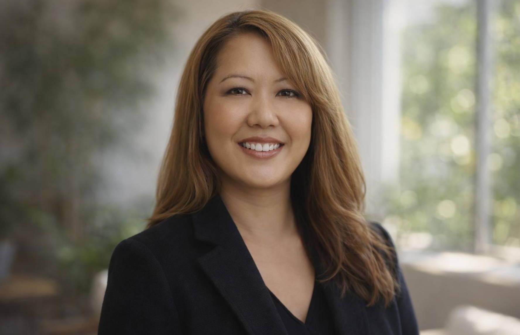 Dr. Jacqueline Ashley, PCC, a Thai-American woman with long brown hair and golden highlights in a black suit, smiling against a softly lit background with light foliage.