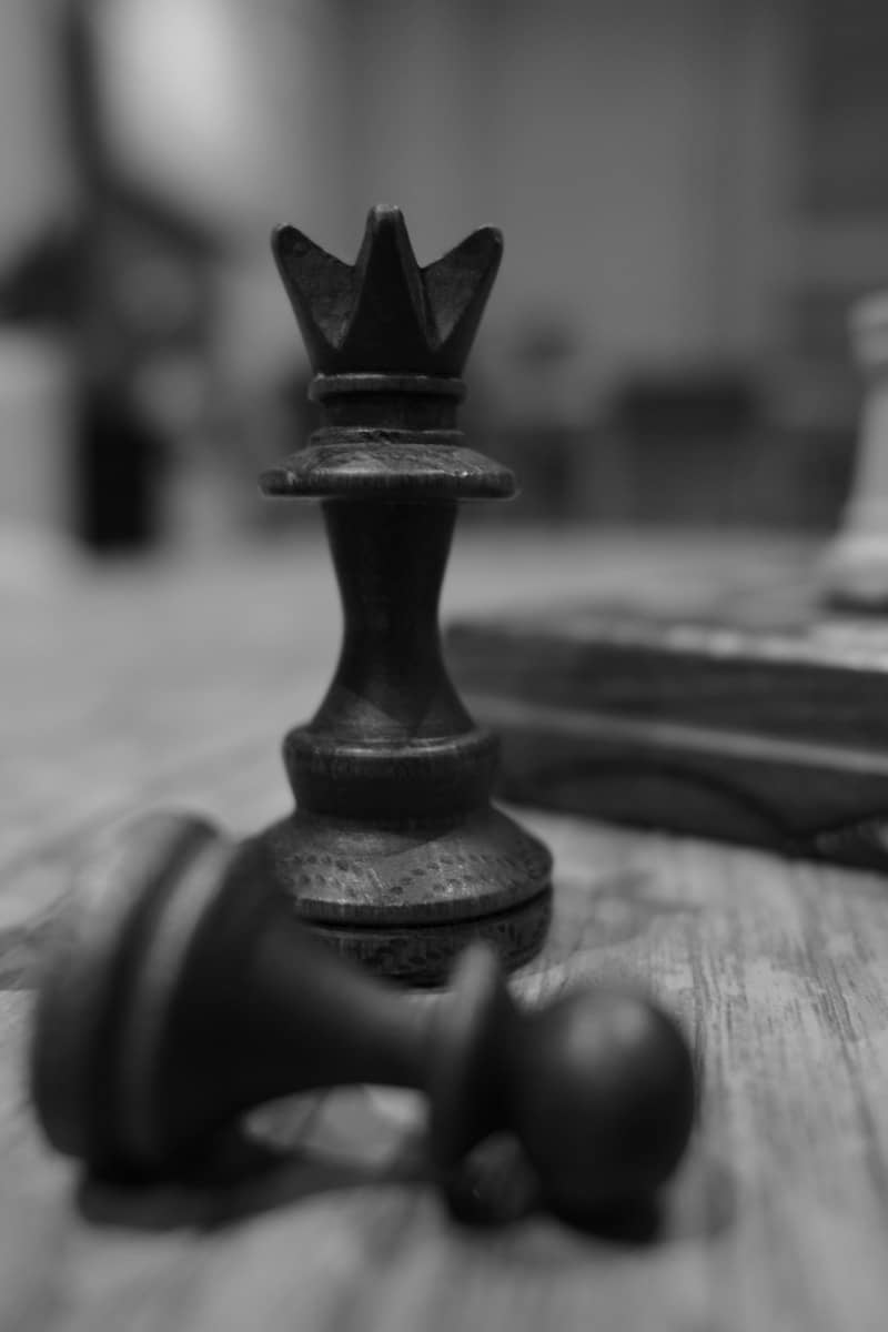 A black chess queen standing behind a fallen black pawn on a wooden board