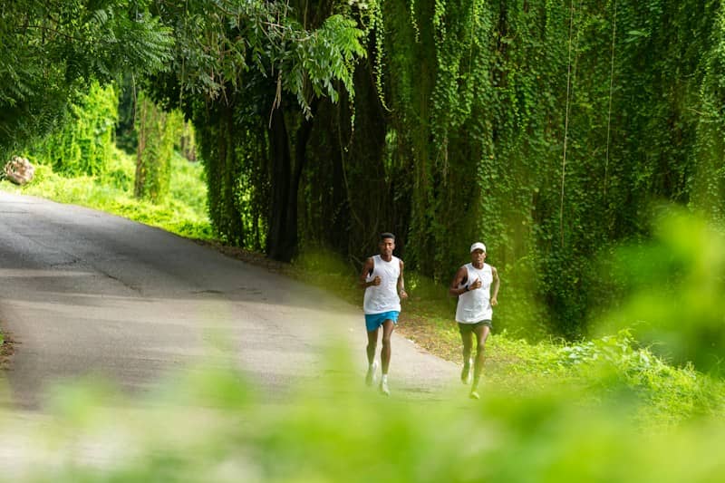 Two men running on a paved road lined with lush green hanging vines