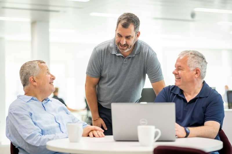Three men talking around a laptop on a white table in an office