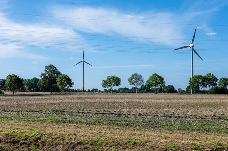 Two wind turbines in a harvested field with trees under a blue sky