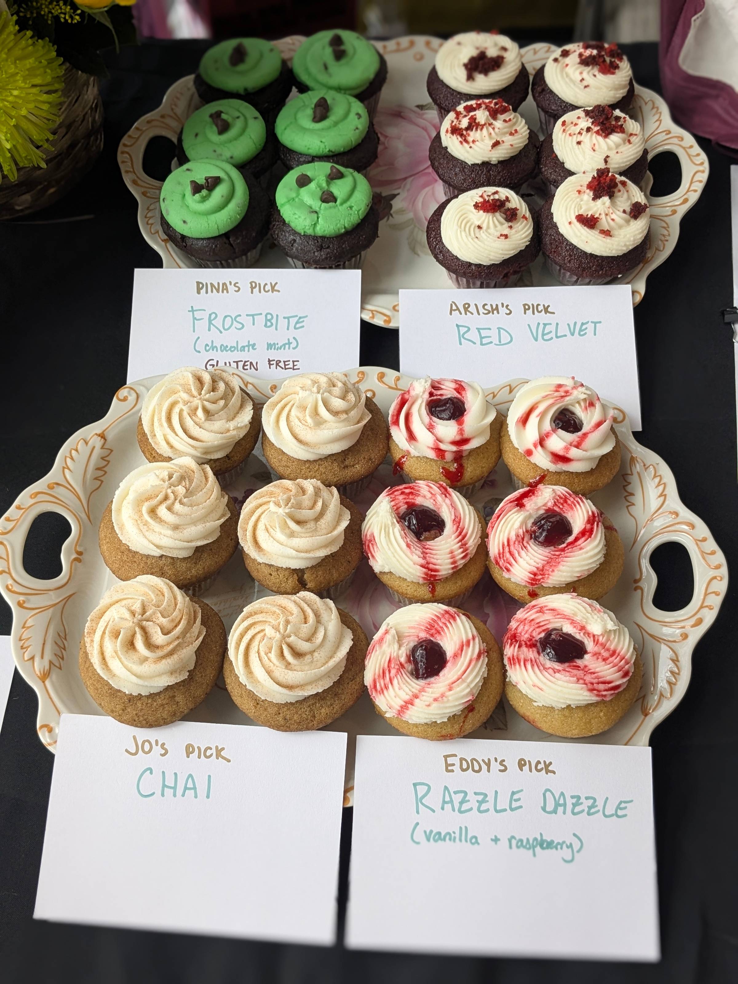 A set of cupcakes with signs. Clockwise, these are: Pina's Pick, Frostbite (chocolate mint, gluten free); Arish's Pick, red velvet; Jo's pick, chai; Eddy's pick, Razzle Dazzle (vanilla + raspberry)