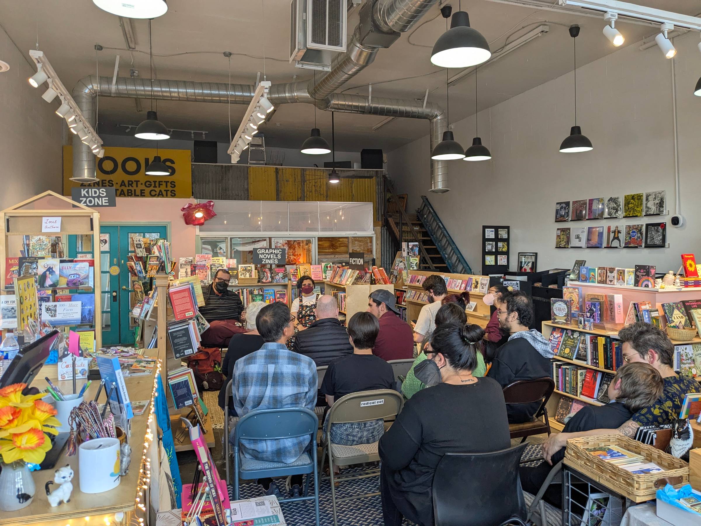 Audience sit in folding chairs in the middle of a bookstore, with the speakers (Naseem and Griffin) in front. Bookshelves box them in.