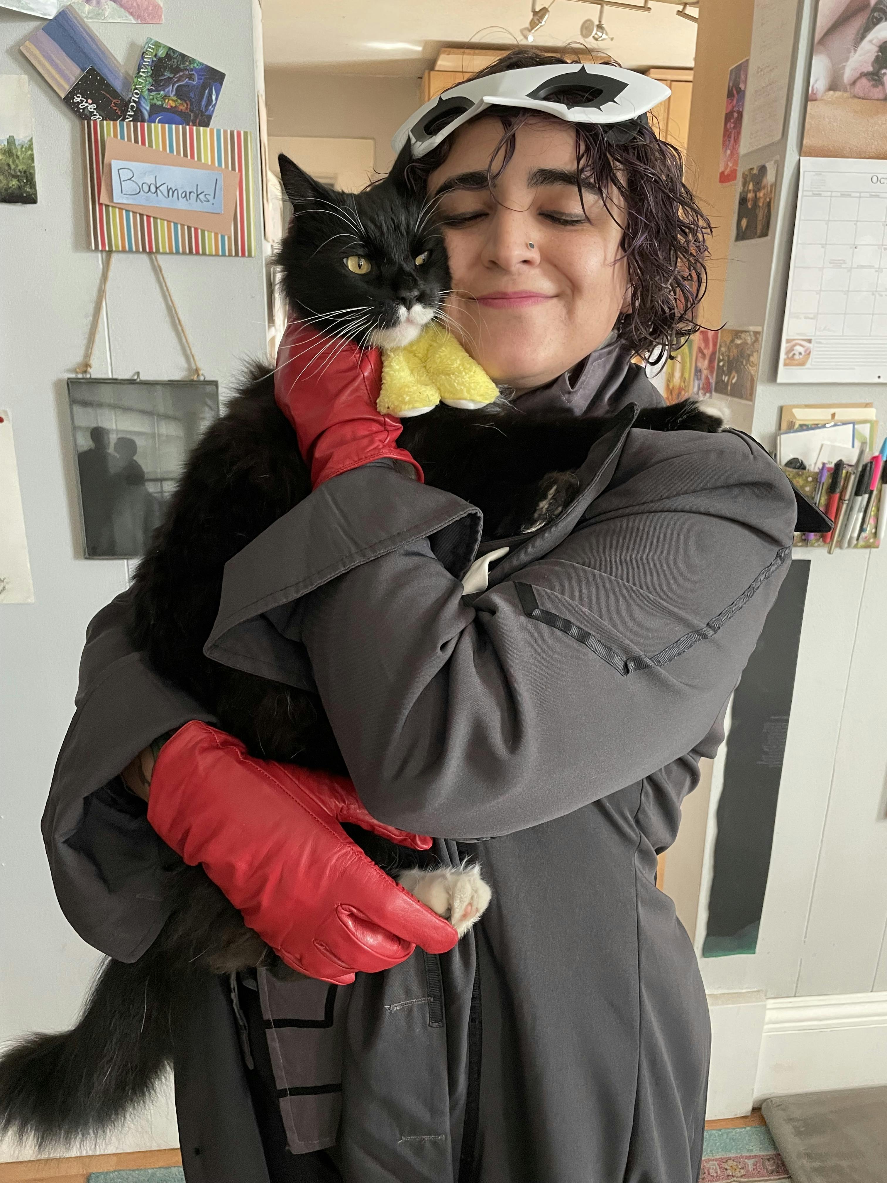 A picture of Naseem dressed as Joker from Persona 5, holding their tuxedo cat Astra.