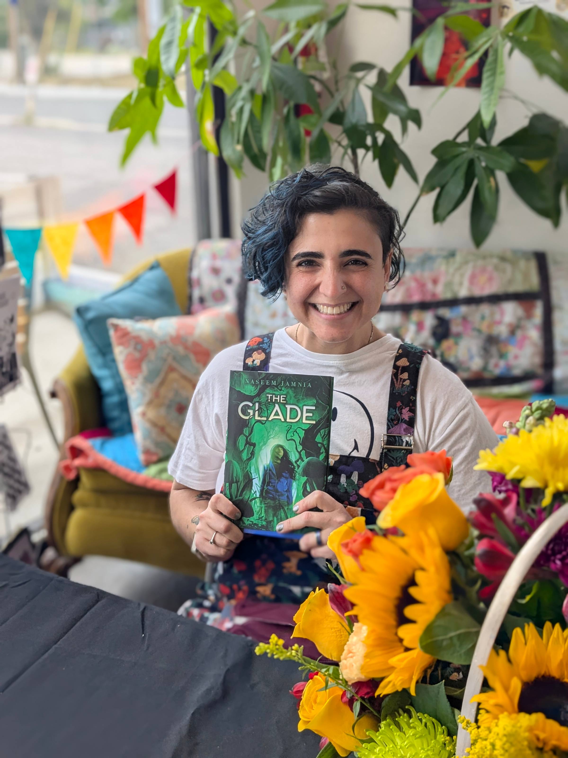 Naseem holds up a copy of The Glade, looking at the camera and smiling. Flowers are in the corner of the picture.