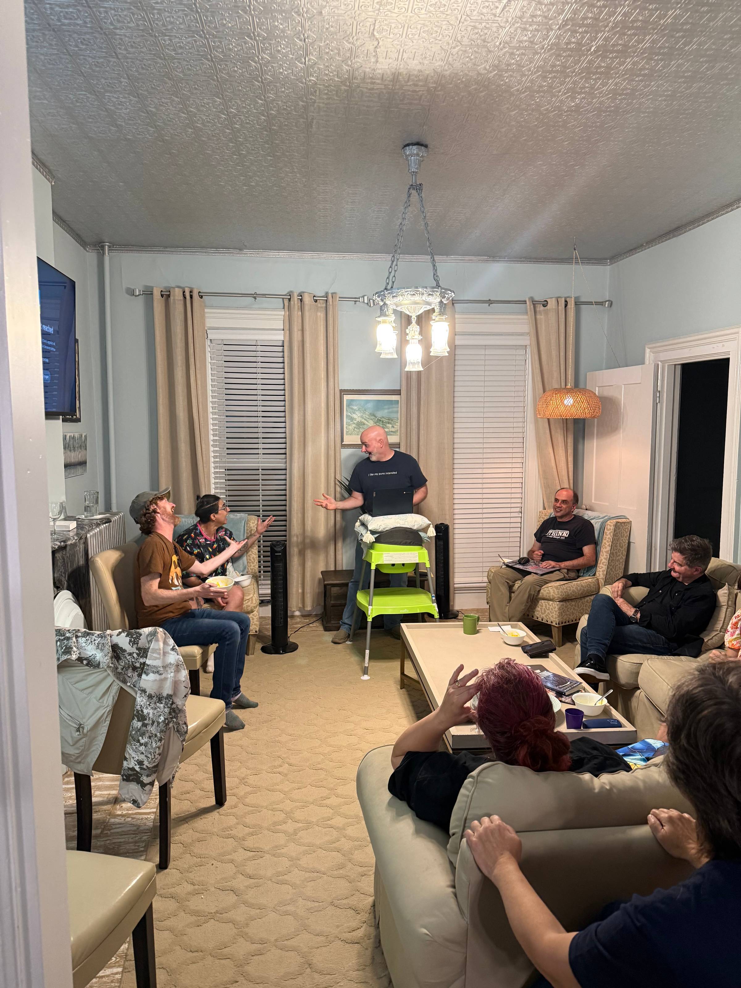 A photo in a living room of author Dan Stout reading, hand outstretched to Naseem, sitting on their left. Naseem, and author Andy Stewart sitting in a seat near them, both extend a hand back to Dan. There are others in the room as well.