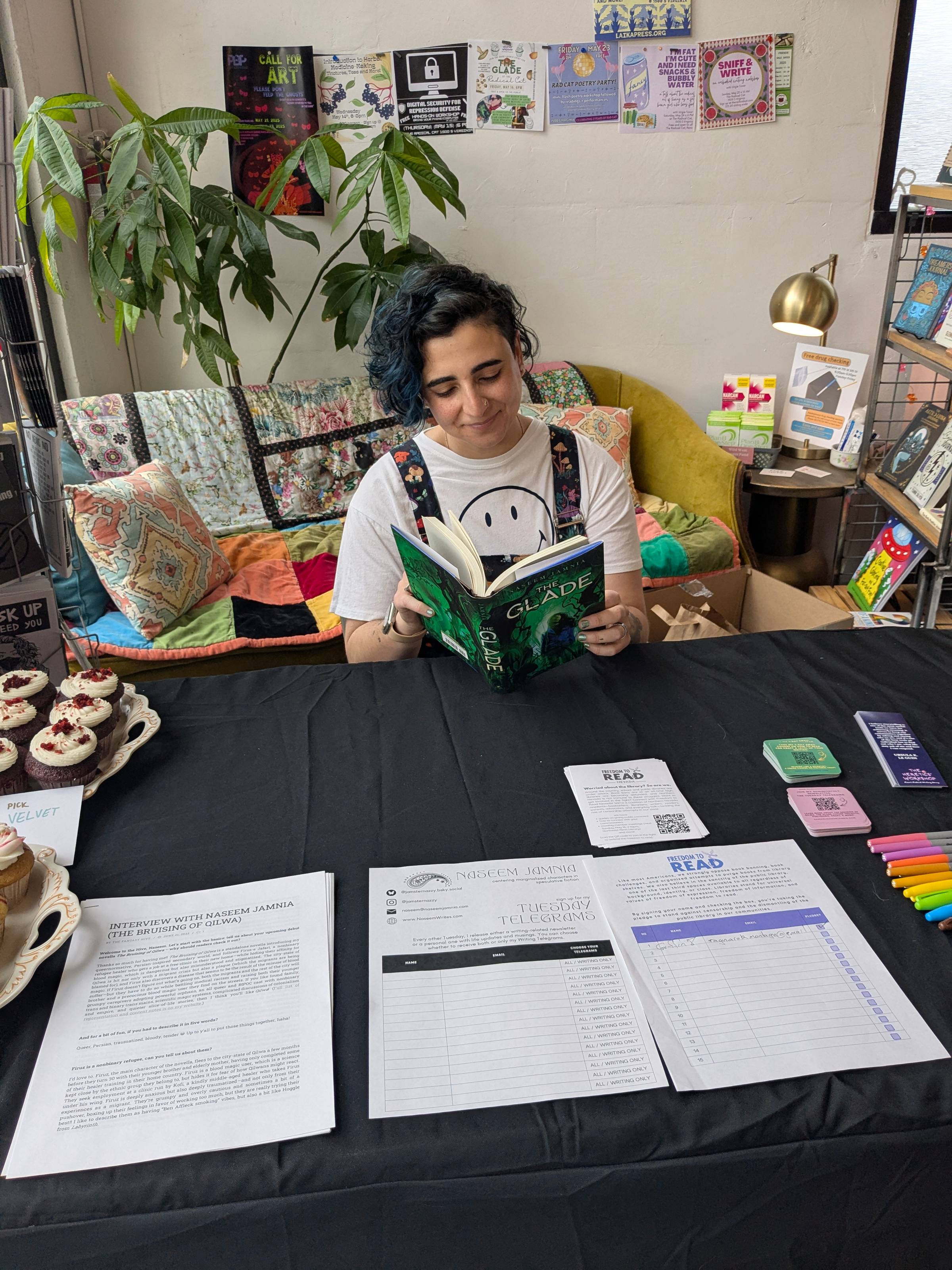 Naseem sits at a table, reading a copy of The Glade. There are sign up sheets  in front of them on the black tablecloth.