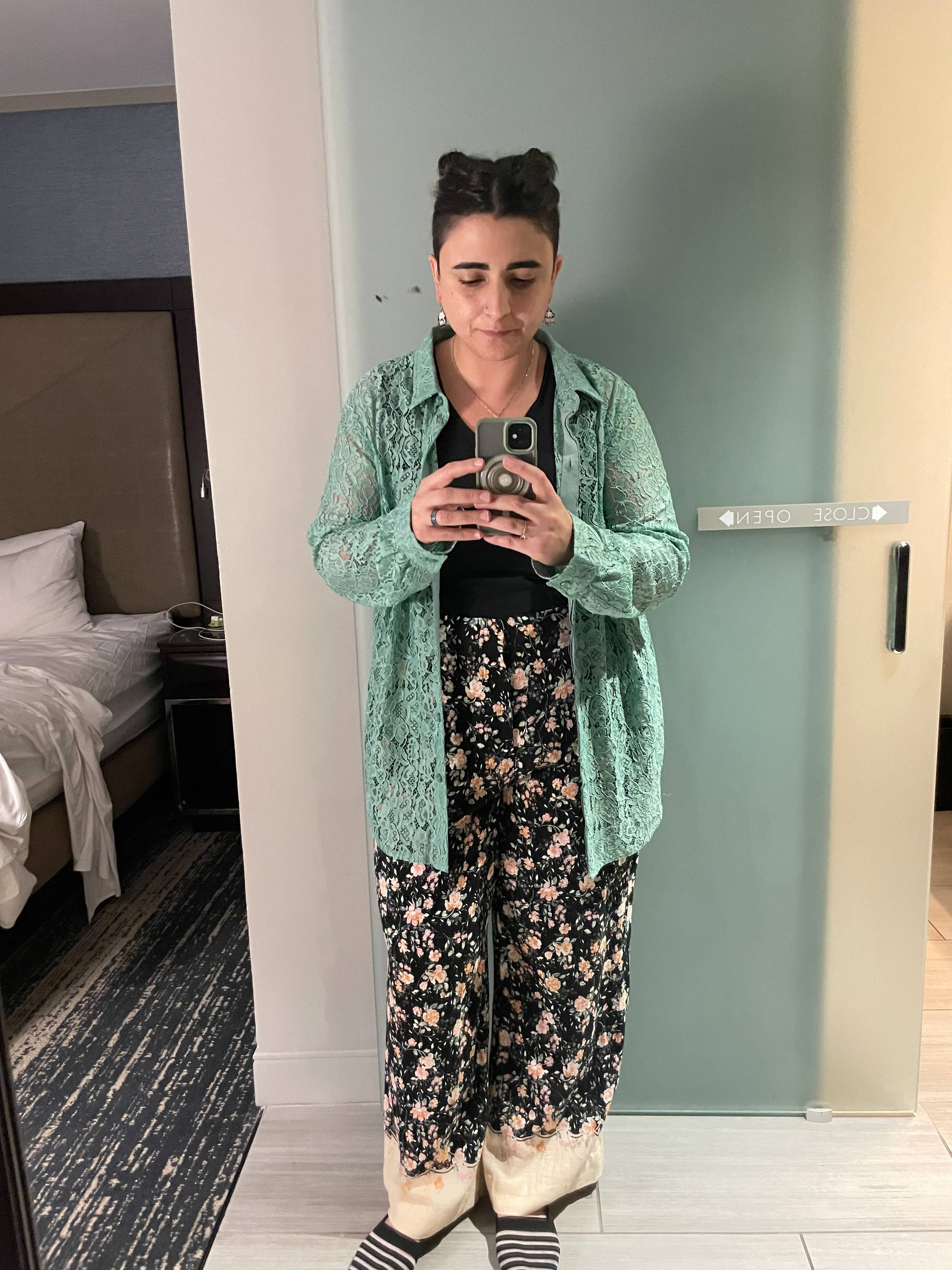 Naseem stands in front of a mirror in a hotel room, taking a picture. They're wearing a black tank, open lacy aqua long-sleeved button down, and black slacks with pink flowers on it and a cream hem. Their hair is in space buns and they wear black flats wi