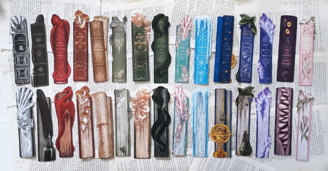 15 fantasy bookmarks spread out showing either side. they're all double sided with the spine on one side and book pages on the other, with little objects growing on or around them, arranged in rainbow order. the themes are: necromancy, old diary, dragons,