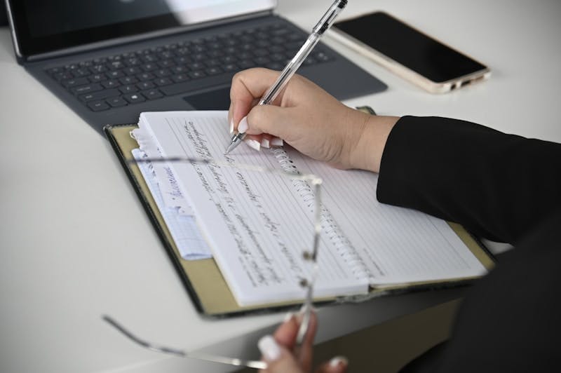 A person writing on a notebook with a laptop in the background