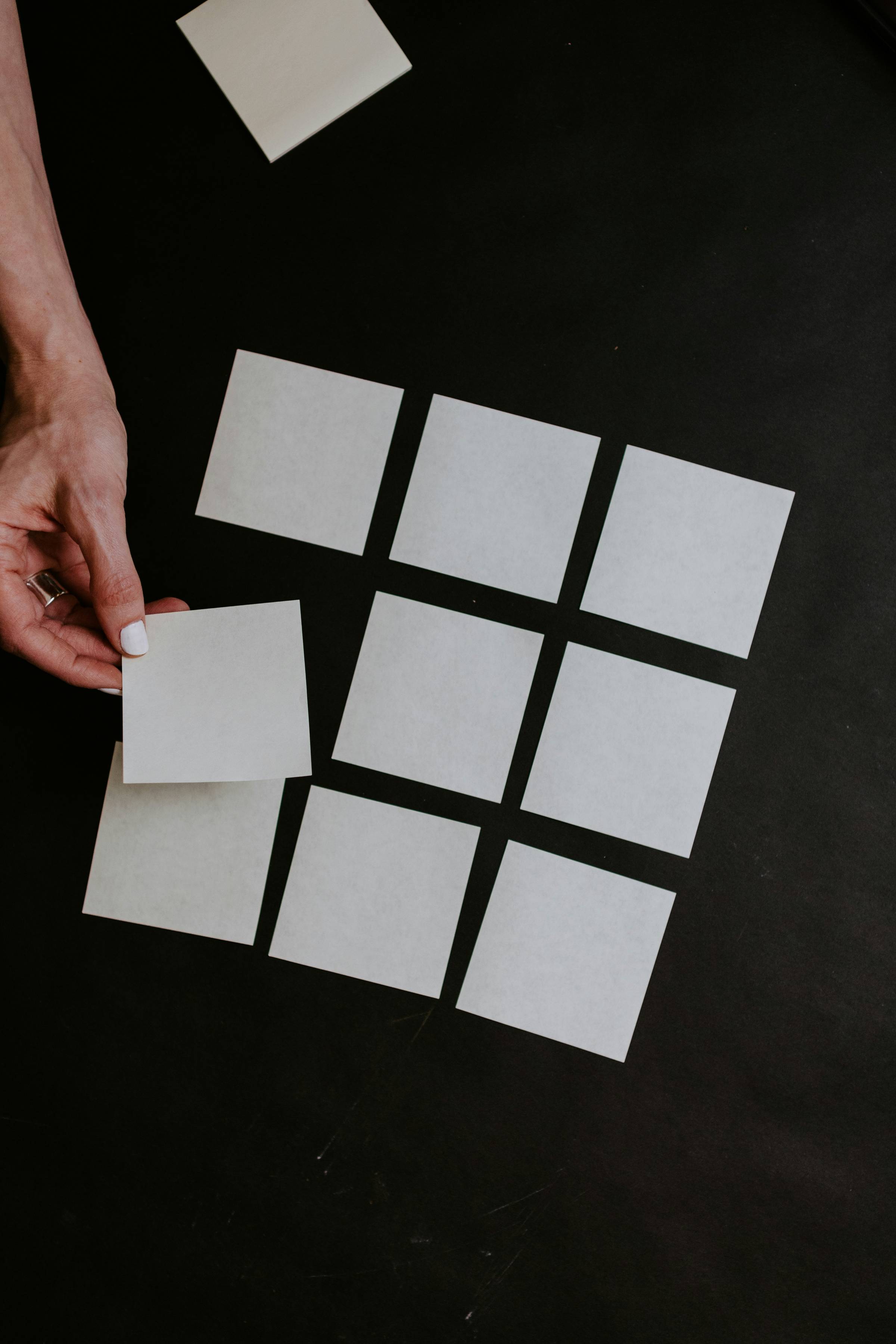 A hand places a blank square sticky note beside a grid of nine other blank sticky notes arranged neatly on a dark surface.