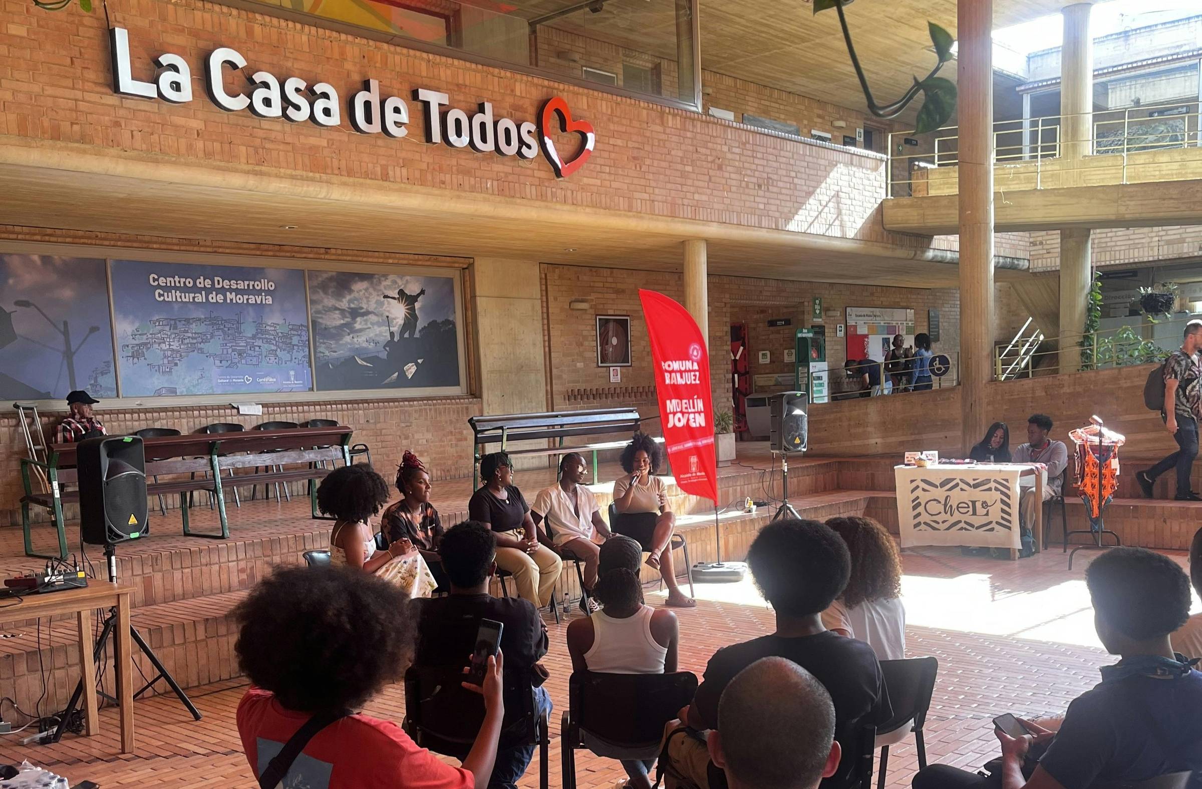 Youth and community members gather for a public forum at La Casa de Todos in Medellín, Colombia, seated in a sunlit courtyard beneath a red canopy.