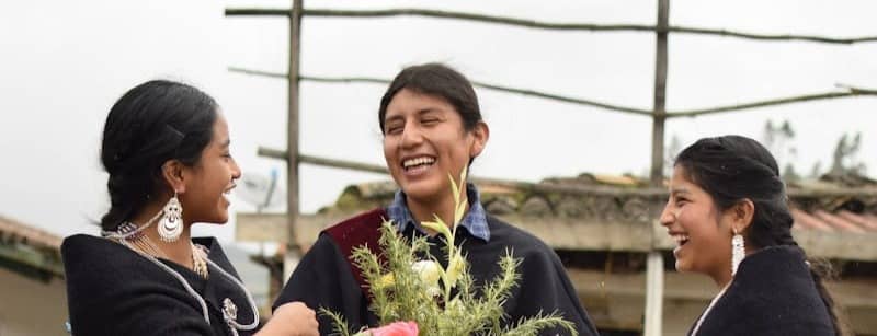 Three Indigenous people in traditional clothing share a joyful moment while holding flowers during a cultural gathering outdoors.
