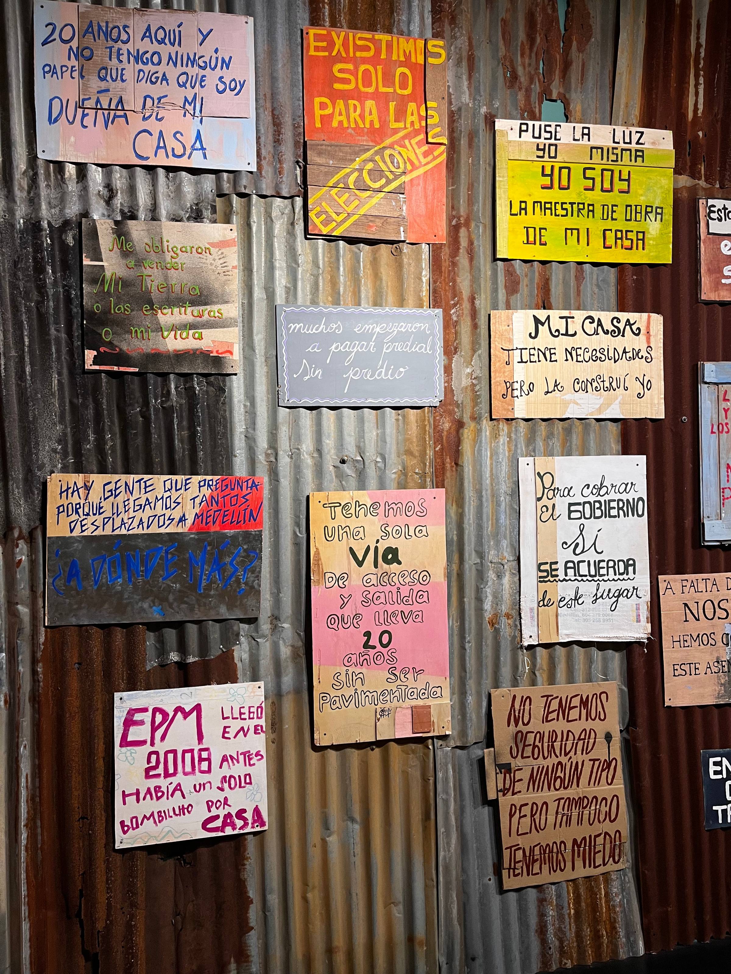 Hand-painted protest signs from displaced residents in Medellín displayed on a corrugated wall at Casa de la Memoria, a museum of collective memory.