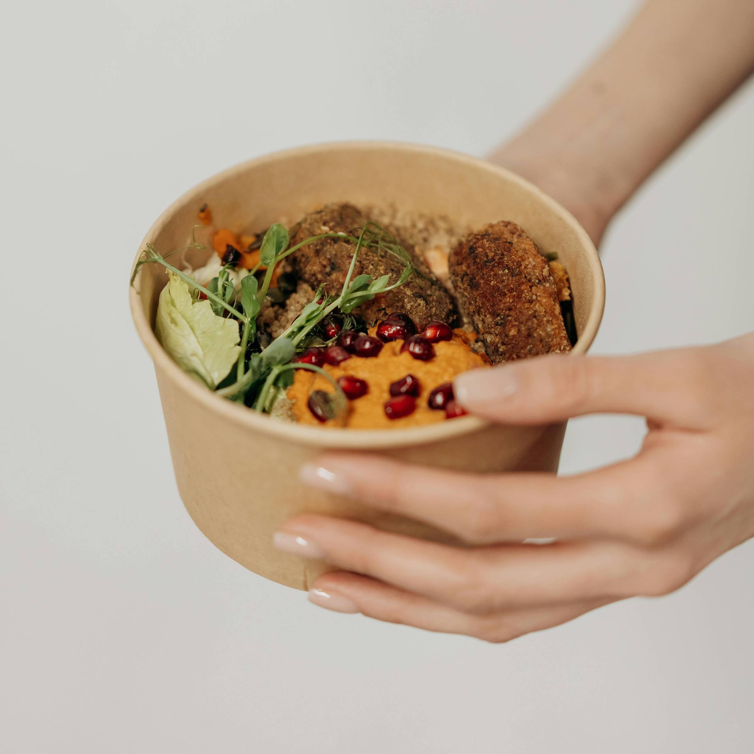 A person holding a cardboard bowl filled with various foods, including fresh greens, falafel, and a topping of orange-colored hummus garnished with pomegranate seeds. The focus is on the hands and the container, set against a plain background.