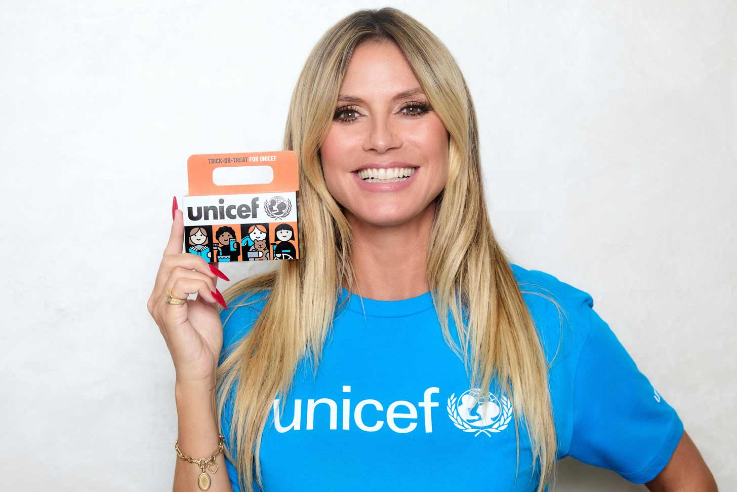 Heidi Klum smiling, wearing a blue UNICEF shirt, holding an orange UNICEF collection box