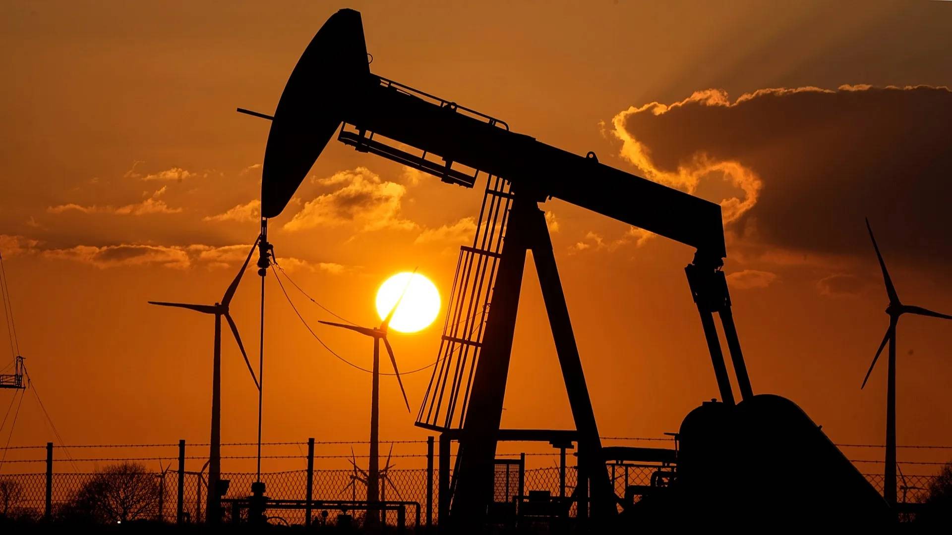 Oil pump jack silhouetted against an orange sunset sky with sun visible behind it; wind turbines visible in the background on either side.