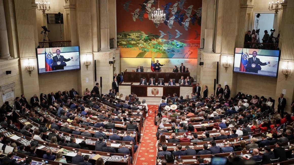 A wide, slightly aerial photo of a room filled with people in rows, several people are at a large desk in the front of the room, one person is speaking at a podium