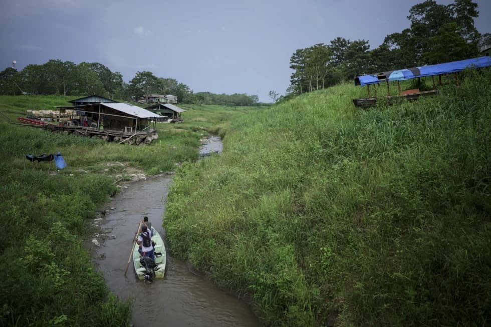 People paddling a boat down a small river with makeshift homes on the banks