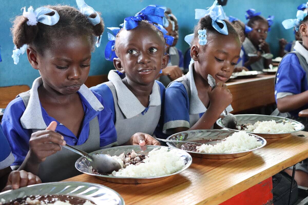 Young girls in blue and grey uniforms sitting at a wood table eating rice and beans