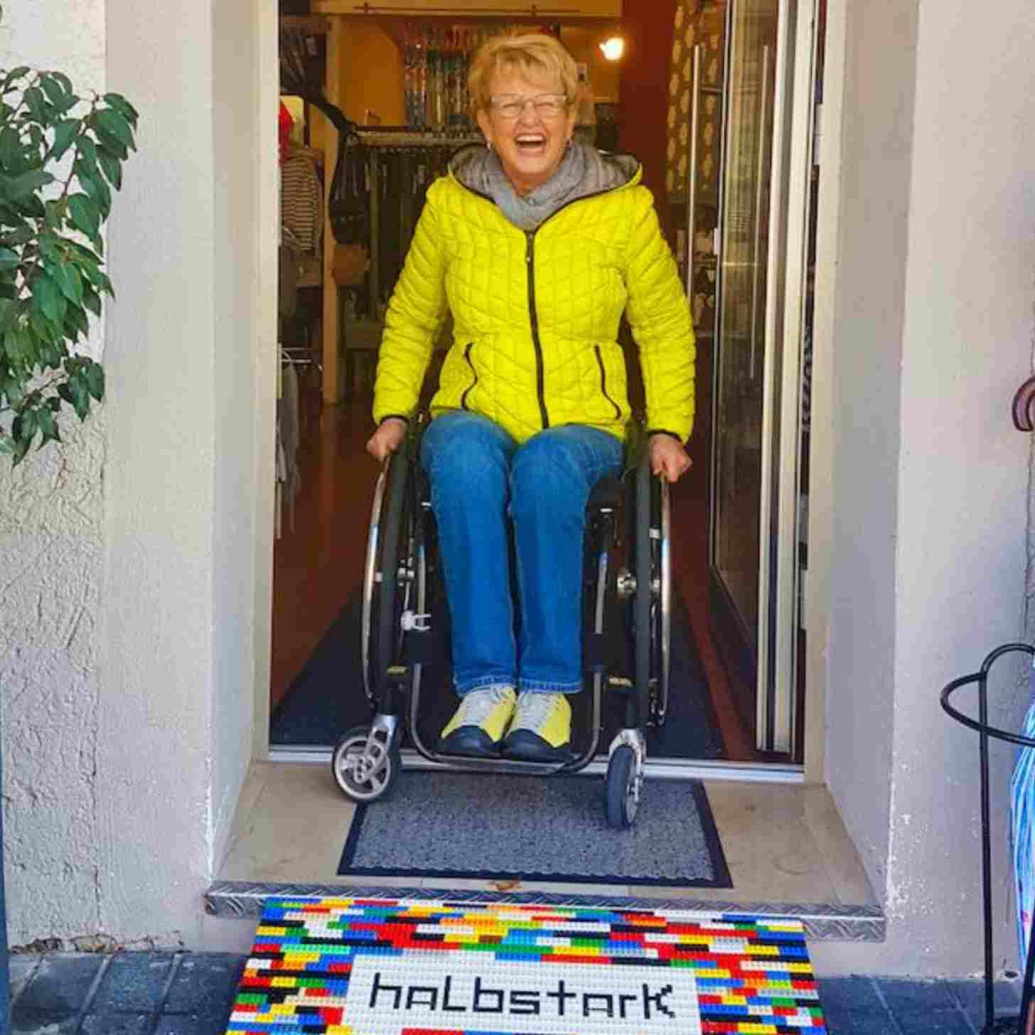 Rita Ebel in a yellow jacket, jeans, sitting in a wheelchair about to go down a ramp made of colorful lego