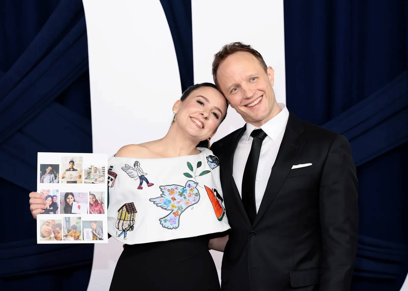 Ms. Rachel and her husband on the red carpet, she's holding a paper with photos of children and their drawings