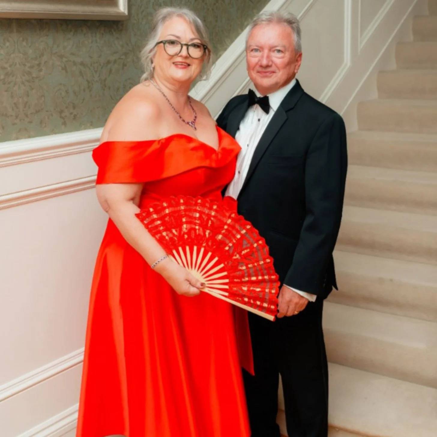 A woman in a red off-shoulder gown holds a red decorative fan while standing next to a man in a black tuxedo with bow tie in an elegant hallway.