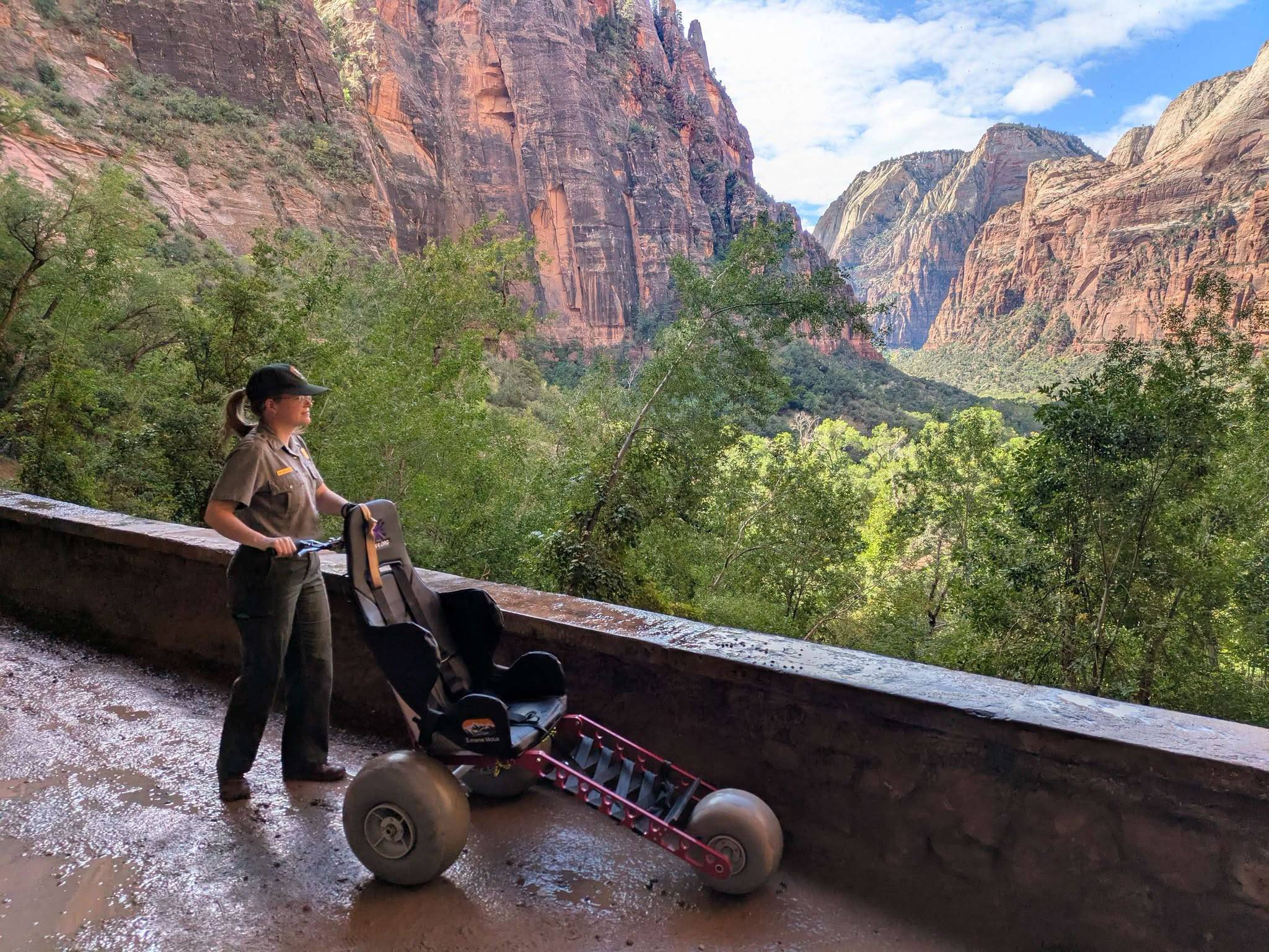 A park ranger stands beside a red all-terrain stroller at an overlook, gazing toward towering red rock canyon walls with green vegetation in the valley below under a partly cloudy sky.