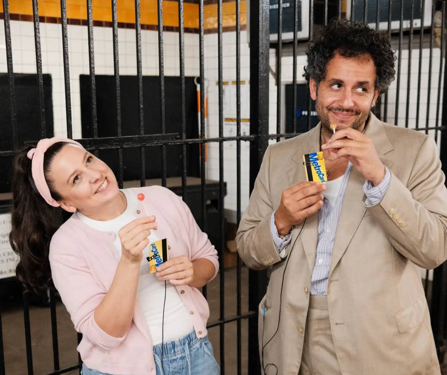 Two people stand in front of metal cages holding yellow and blue packaged snack bars. The woman on the left wears a pink headband, pink cardigan, and jeans. The man on the right wears a beige suit jacket and light blue shirt.