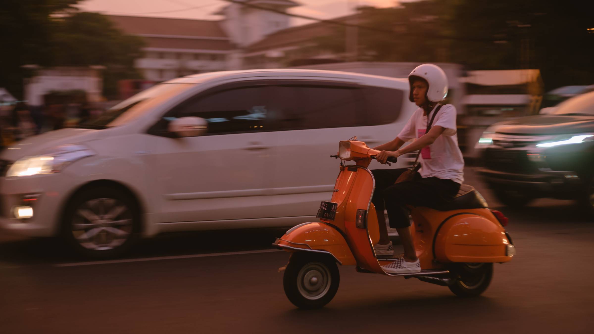 A person driving an orange moped next to a white car