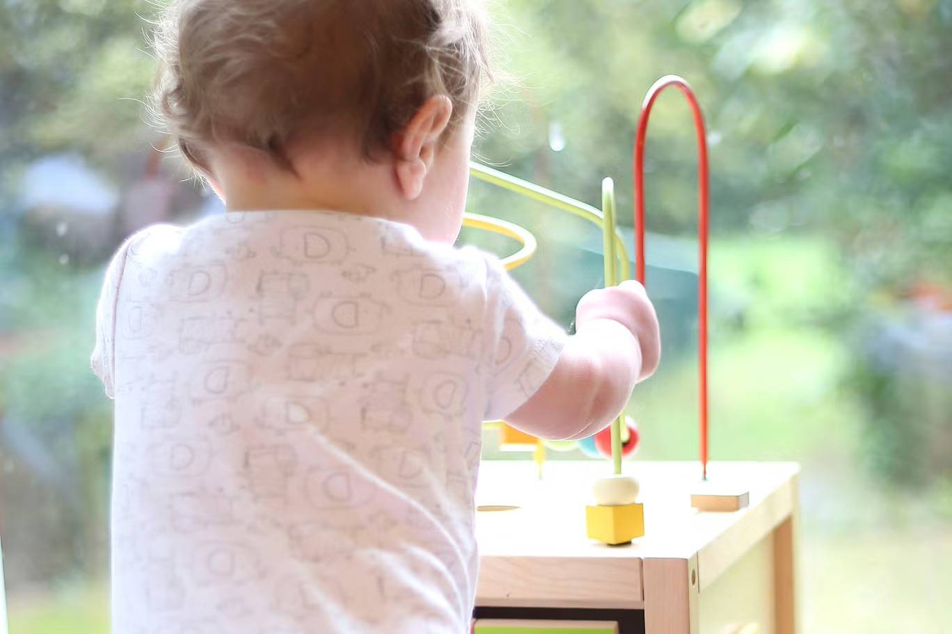 A baby playing with a table-top toy pictured from behind wearing a white onesie with line drawings on it