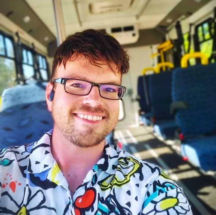 Person in a colorful shirt and glasses taking a selfie at the front of a bus