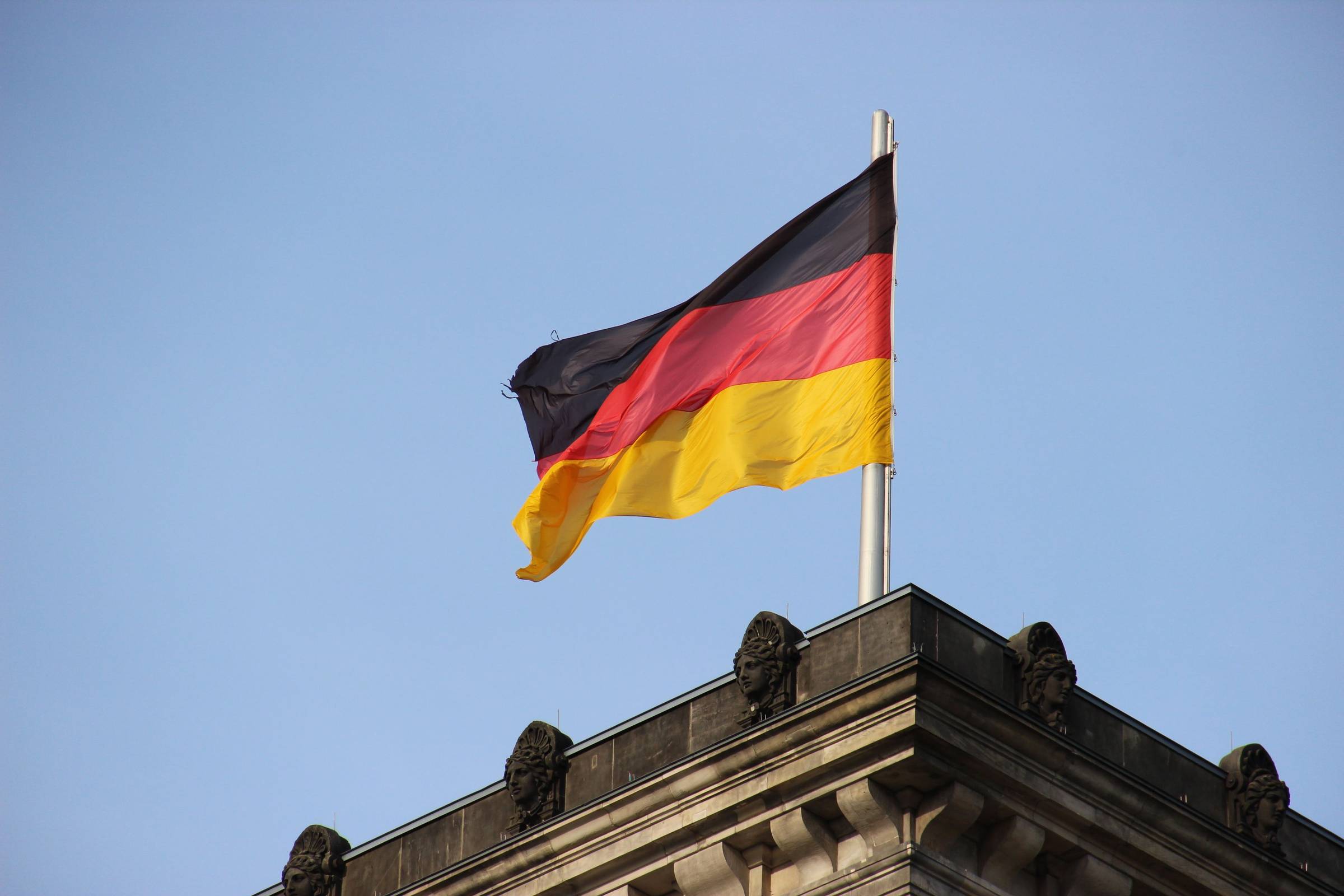 The German flag waves against a clear blue sky, mounted on a pole above an ornate stone building with decorative sculptural elements.