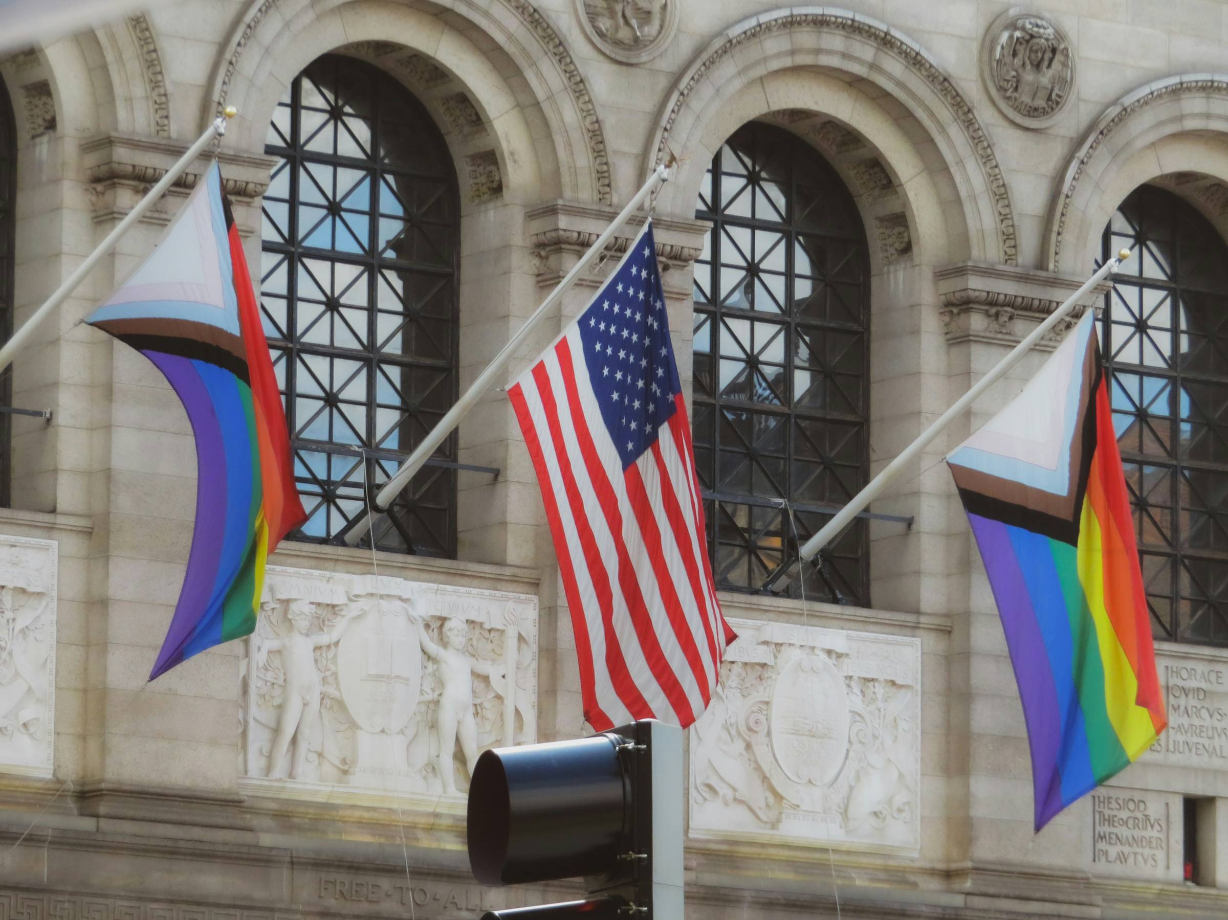 Two progress pride flags on either side of an American flag on the outside of a building